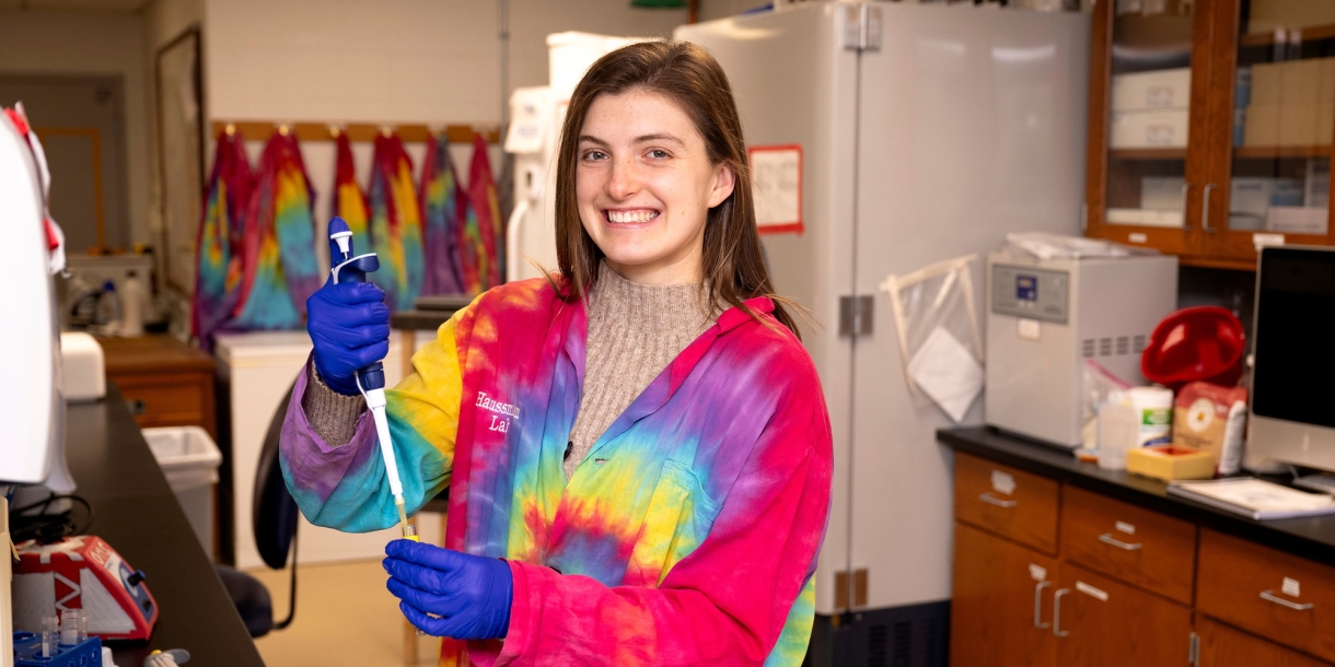 Meghan Catherwood '25 wears a bright, multi-colored tie-dyed lab coat and blue gloves while smiling and holding a pipette in a lab on campus.