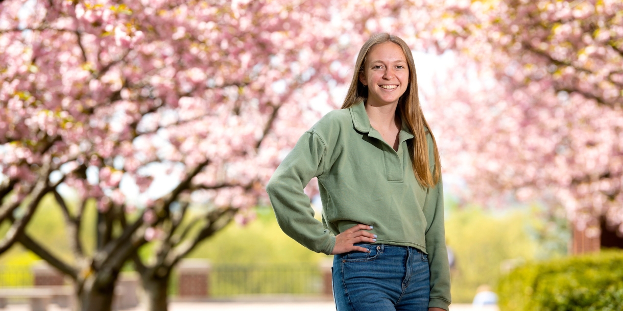 Student Ally Clarke poses to the left-center of the frame standing between two rows of pink cherry blossom trees in full bloom