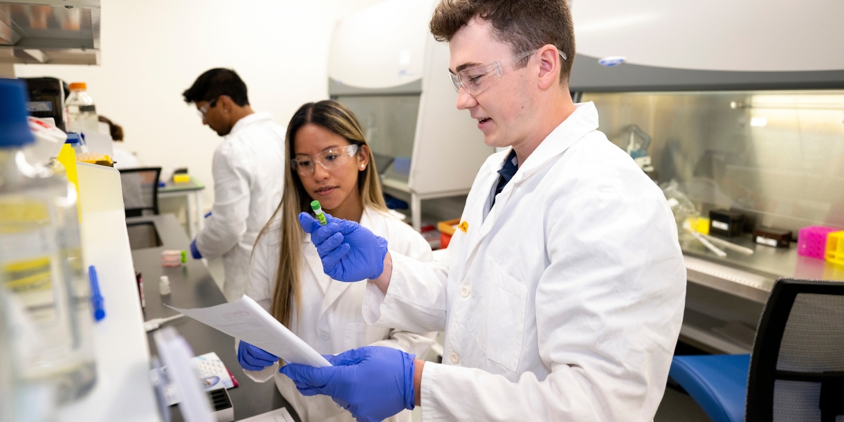 Professor Olivia Boerman and Connor Kozick &#039;26 are wearing white lab coats and purple gloves in a lab setting.