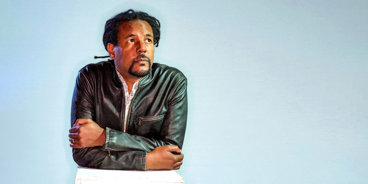 Portrait of author Colson Whitehead leaning forward against a white box, staring upward and off camera to the right.