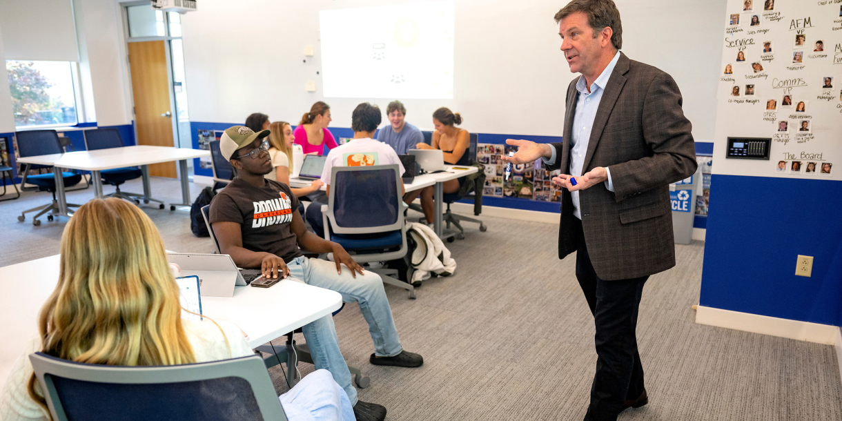 A professor in a suit jacket stands in front of a white board with students surrounding him in a classroom.