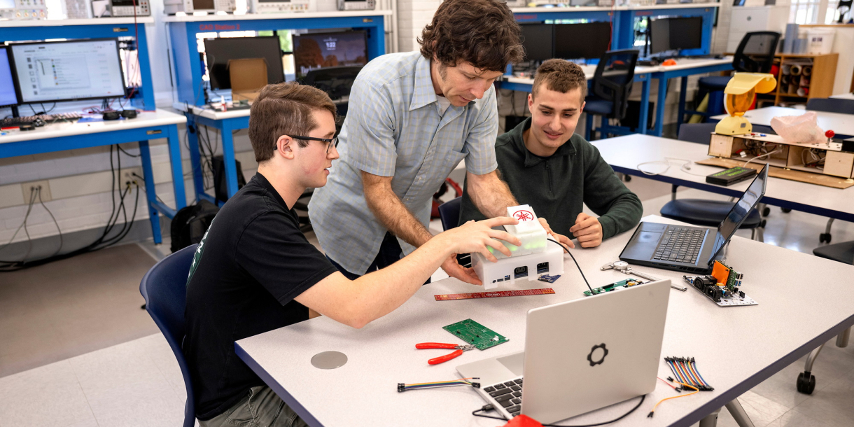 Two students sit at a table in a makerspace with a staff member in between them as they look at pieces of technical hardware together.