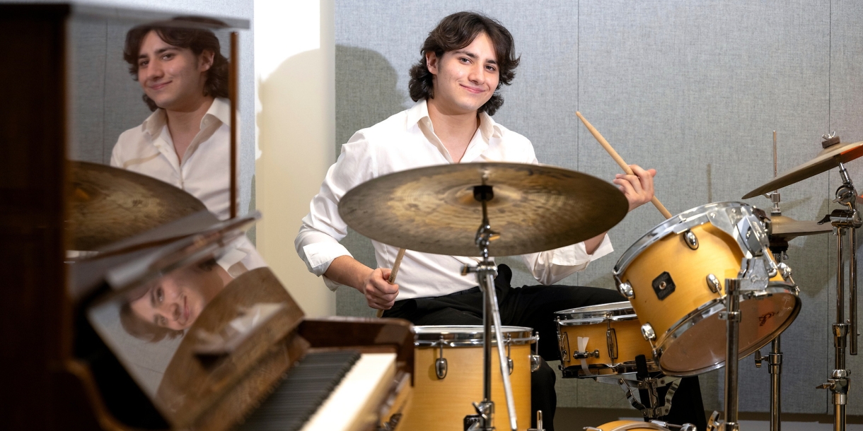 Darren Mayrowetz '27, dresed in a white collared shirt and black pants, plays a drum set next to a piano.