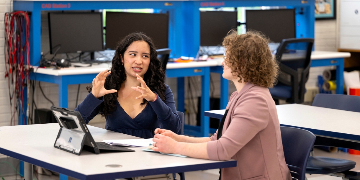Aura Chuck Hernandez sits at a table conversing with Professor Annie Ross.
