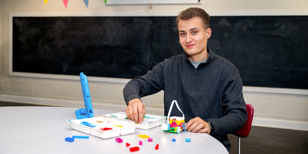 Aiden Cherniske '27 sits at a table and smiles with colorful components of robotics materials in front of him. 