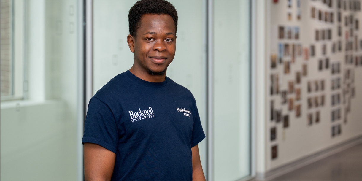 Emanneul Mudungwe stands and smiles in a hallway in Holmes Hall while wearing a navy Bucknell T-shirt