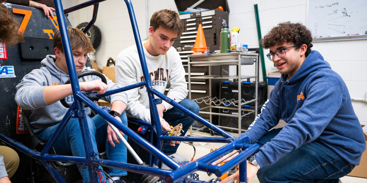 A group of students gather around the frame of a stripped down vehicle.