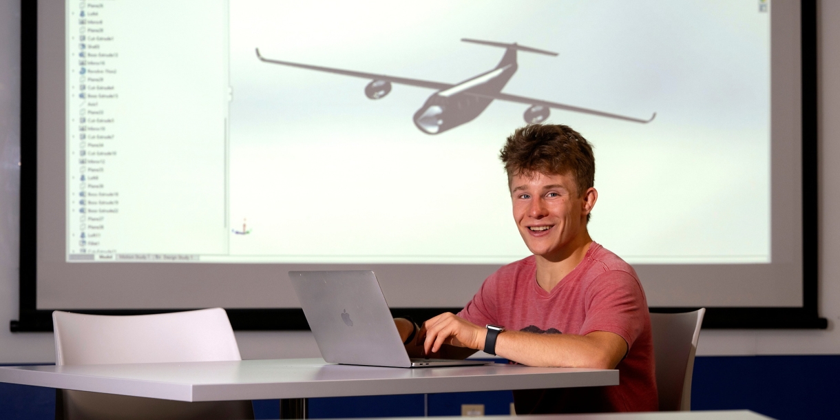 Kade Davidheiser sits at a table on his laptop with an image of an airplane model projected on the screen behind him