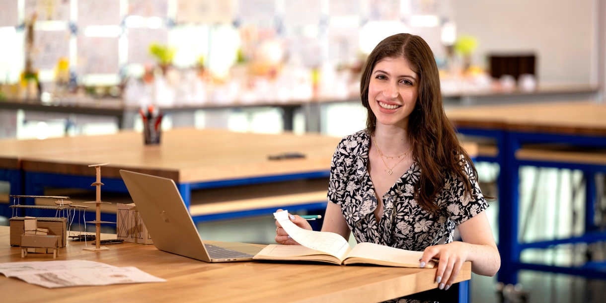 Katie Raso '28 sits in a classroom at a desk with a laptop and papers in front of her and smiles.