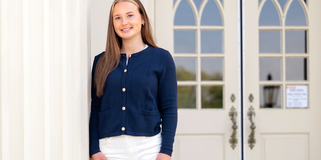 Madelyn Hudak wears a blue top and white pants and leans slightly against a white column outside a building on campus.