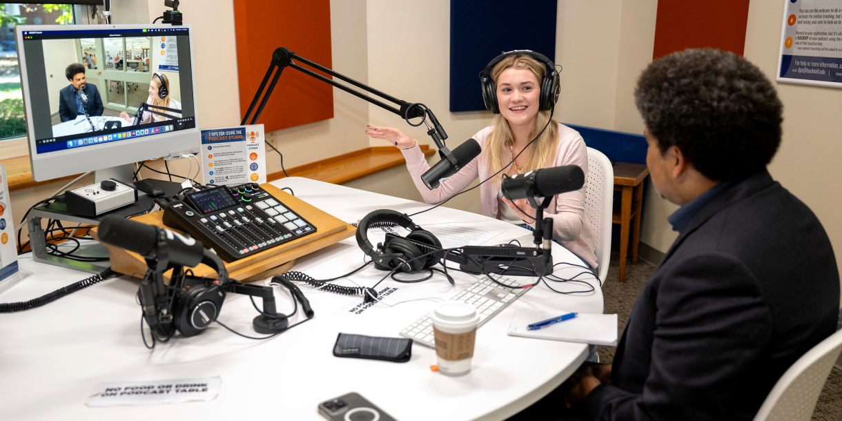 Rayna Tyler '27 wears headphones and sits at a table with a microphone and computer screen while speaking to a guest, who has his back to the camera.