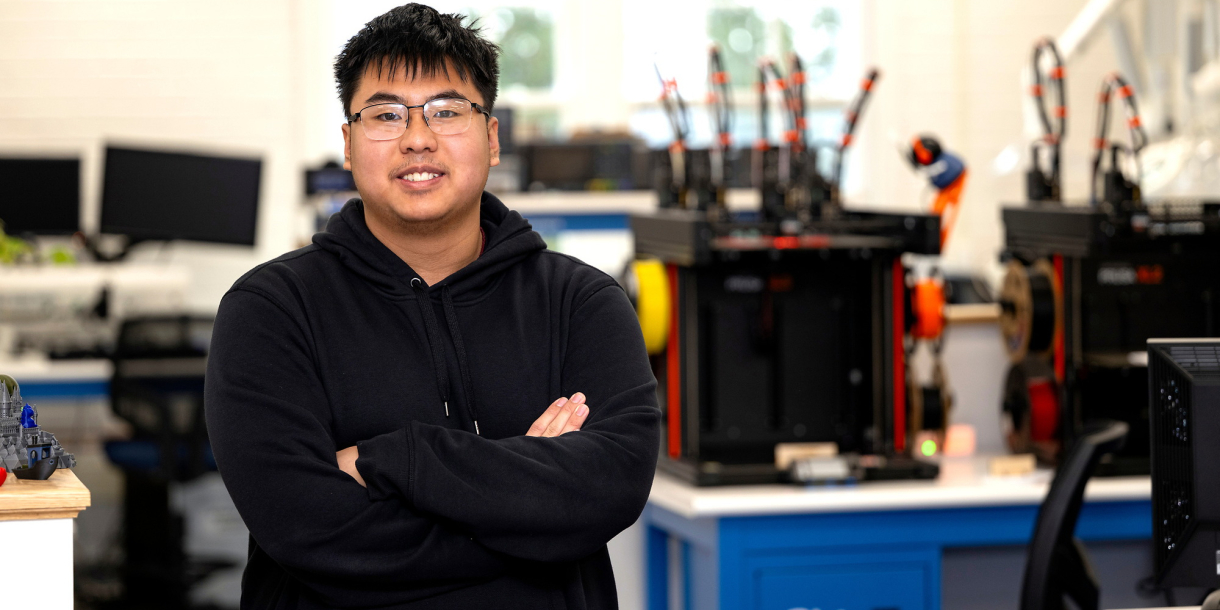 Richard Chi stands in a makerspace and smiles while wearing glasses and a navy hoodie.
