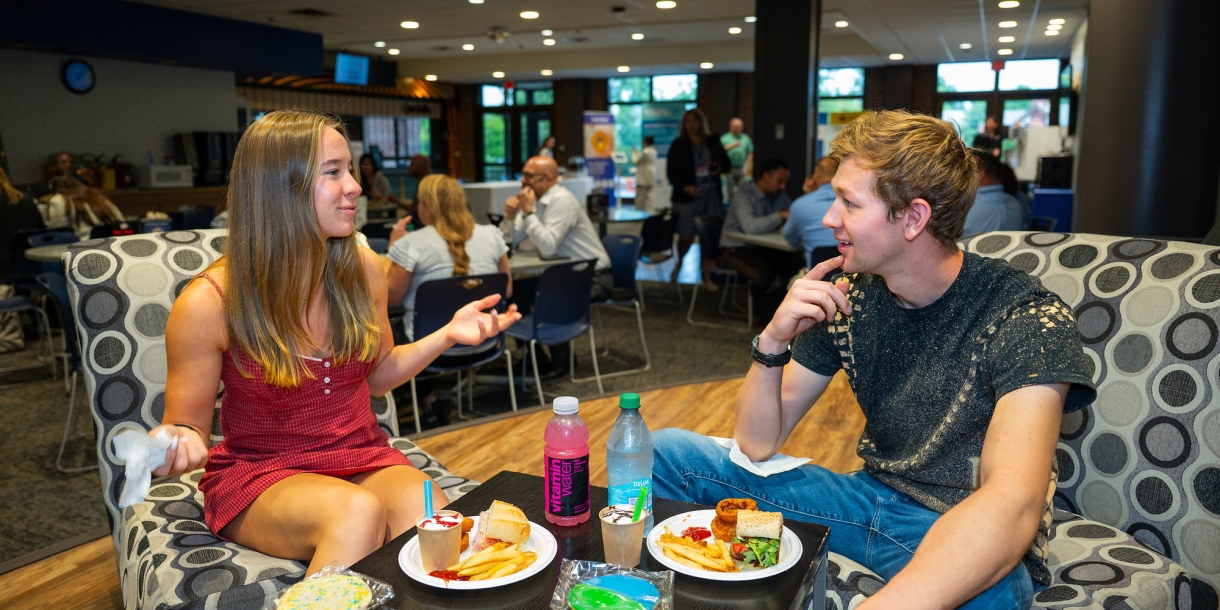 Two students sit in The Bison on colorful chairs and talk while eating food.