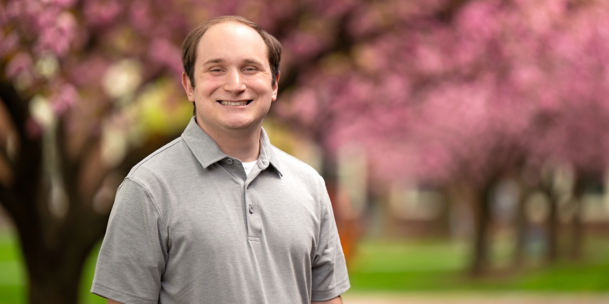 Zane Hensal wears a gray short-sleeved colored shirt and smiles on campus with cherry-blossom trees behind him.