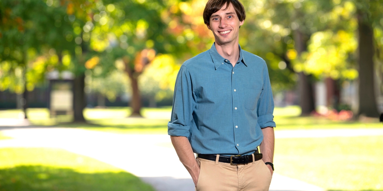 Sam Gutekunst is wearing a blue shirt and tan pants and stands and smiles on campus with sunlight shining through green leaves behind him. 