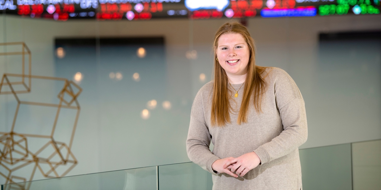 A student stands inside a building with a high-tech ticker behind her, displaying current stock prices. 