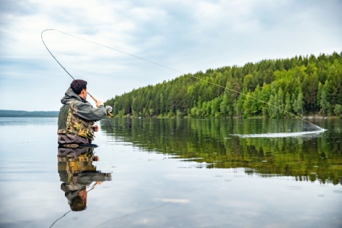 A person in a fishing vest stands in water waist deep while fly fishing.