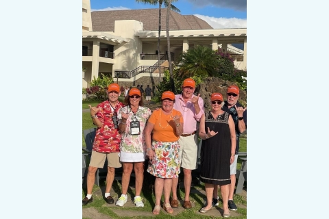 A group of alumni stand in front of a resort building orange hats and giving the &quot;hang 10&quot; hand sign.