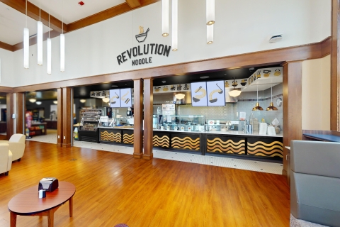 An artist rendering of an Asian takeout restaurant in MacDonald Commons. A sign stating Revolution Noodle appears above a serving counter with instant noodle patterns along the front. A lounge area with leather chairs is in the foreground.