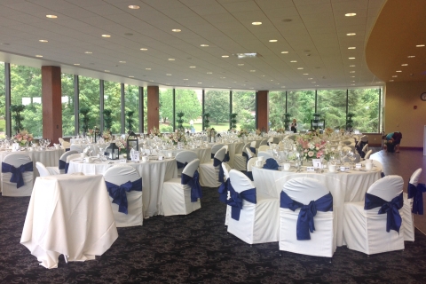 Terrace Room in the Elaine Langone Center setup for a wedding. Tables set with white tablecloths; chairs with white seat coverings and blue bows. Table settings and flower arrangements in place on each table. 