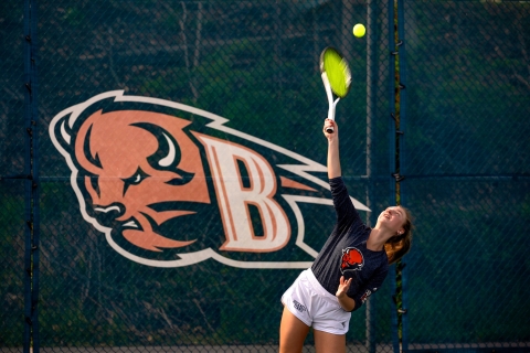 Anna Lajos '25 hits a tennis ball with her racquet in front of a Bucknell Bison decal on a tennis court wall.