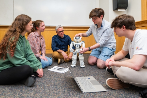 Professor Felipe Perrone and a group of students sit on the floor with a robot and talk.
