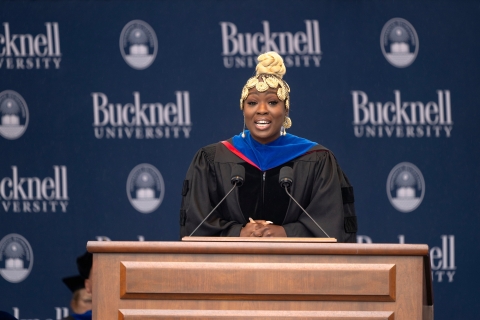 Nadia Sasso &#039;11 speaks on the Commencement stage behind a podium and microphones and in front of a Bucknell University logoed backdrop.