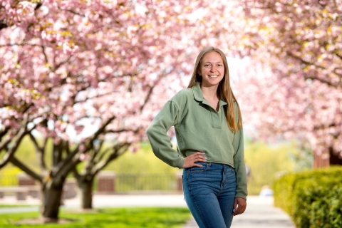 Student Ally Clarke poses to the left-center of the frame standing between two rows of pink cherry blossom trees in full bloom