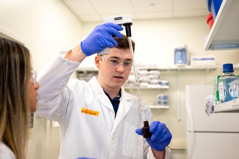 Connor Kozick &#039;26 wears a white lab coat and purple gloves and goggles while holding a piece of lab equipment as Professor Olivia Boerman looks on.