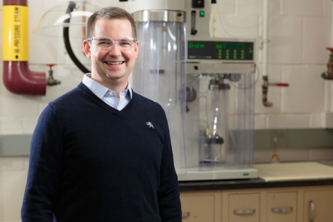 Ryan Snyder is wearing a navy sweater and clear-framed glasses and stands and smiles in a lab with equipment behind him and smiles.