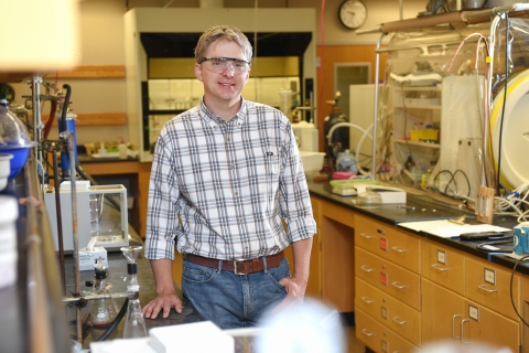 Will Kerber smiles while wearing safety goggles and a white plaid shirt and jeans in a lab