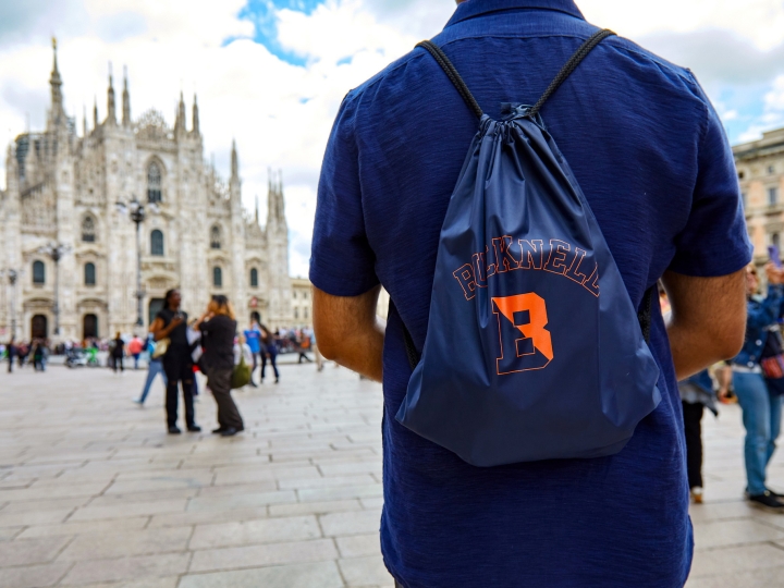 Bucknell student in a square in Milan. Student is facing away from the camera, wearing a Bucknell backpack. Cathedral in background. 