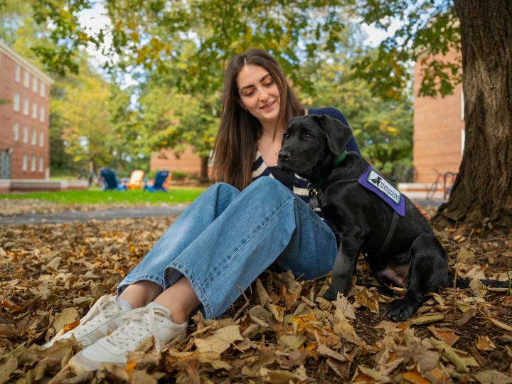 Emma Feld sits on a blanket of leaves on the ground with a young black lab puppy.