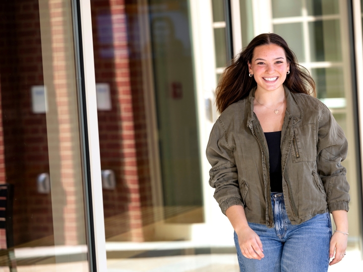 Elizabeth Malley wears a tan jacket and jeans and walks in front of a building on campus with a very big smile.