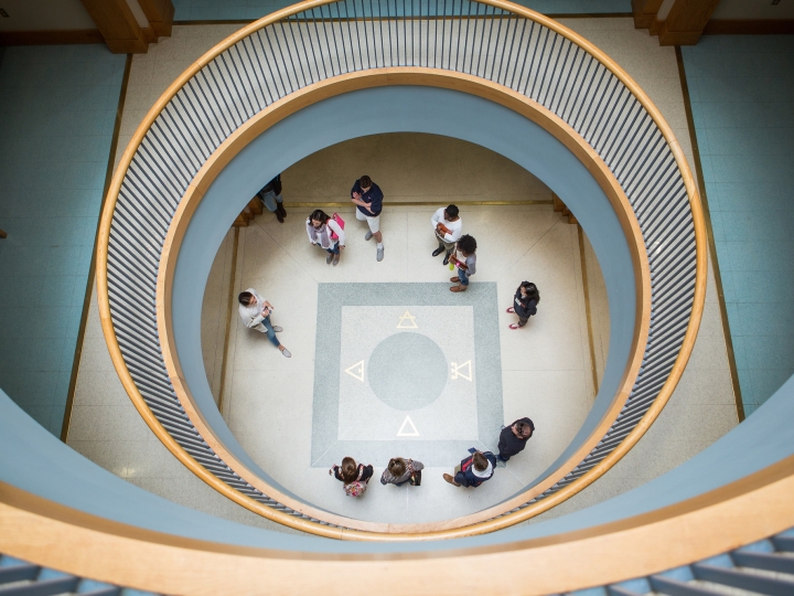 Overhead view of a spiral staircase with people standing in a circle on the ground floor. The stairway's curves frame the scene, creating a concentric pattern.