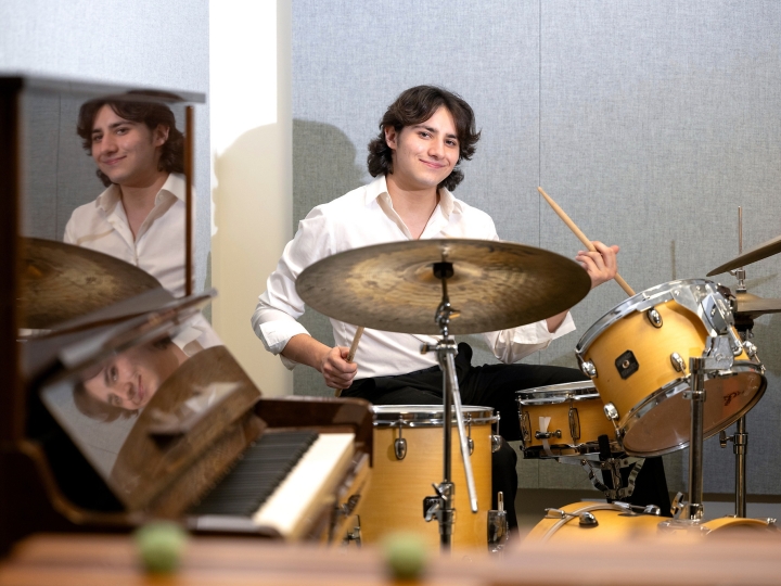 Darren Mayrowetz '27, dresed in a white collared shirt and black pants, plays a drum set next to a piano.