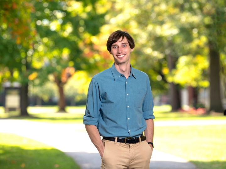 Sam Gutekunst is wearing a blue shirt and tan pants and stands and smiles on campus with sunlight shining through green leaves behind him. 