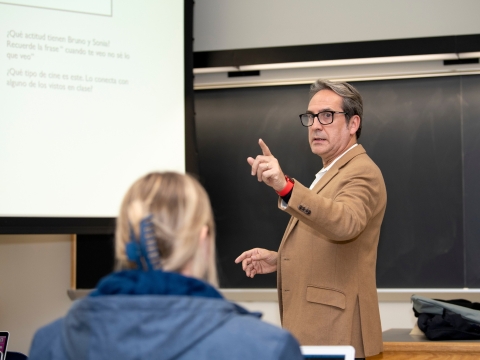 Professor Fernando Blanco stands in front of a chalkboard and projector screen, raising a finger as he makes a point during a class lecture