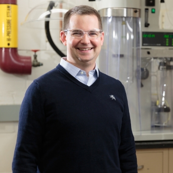 Ryan Snyder is wearing a navy sweater and clear-framed glasses and stands and smiles in a lab with equipment behind him and smiles.