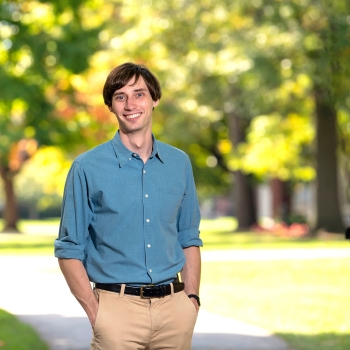 Sam Gutekunst is wearing a blue shirt and tan pants and stands and smiles on campus with sunlight shining through green leaves behind him. 